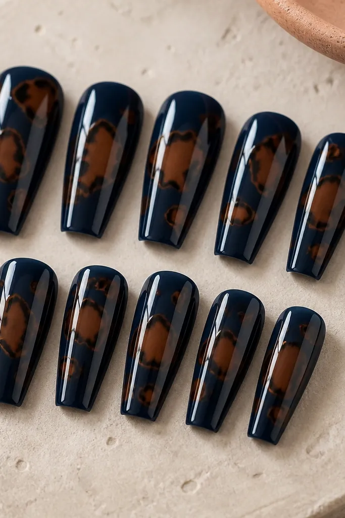 Ten glossy coffin nails. The base is midnight blue. Tortoise shapes appear in cocoa brown with a few bigger, opaque "chunks" of lighter brown near the center of each nail. The pattern has irregular borders that look like shell pieces, not dots.