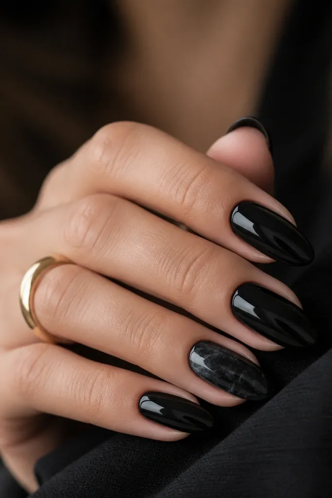 All nails are glossy black except the ring finger, which has black marble swirls. The marble has thin grey-black waves and small white micro-speckles scattered lightly across the surface. The finish is glossy and smooth with a natural stone look.