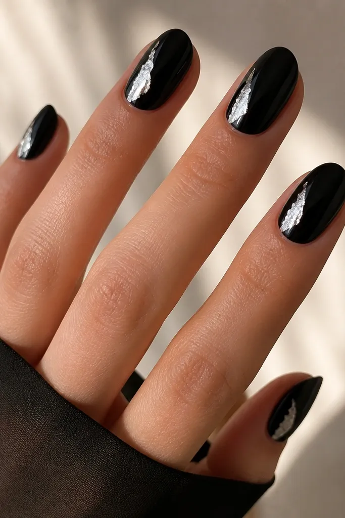 Nails painted glossy black. On each nail there's a silver foil half-moon placed on the left sidewall near the middle, slightly curved and uneven like real foil. The foil catches bright reflections while the rest of the nail stays plain black.