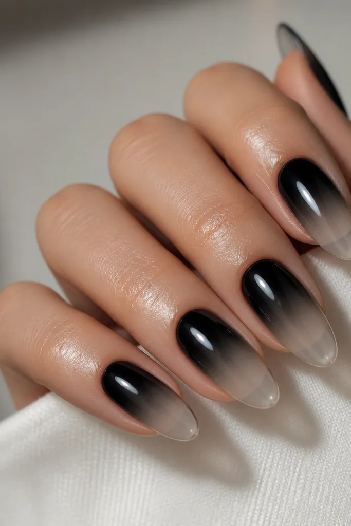Nails with a reverse ombre: the cuticle area is deep black, and the color fades into a clear or sheer tip. The transition is smooth with no harsh line. The nails look glossy and hydrated like a gel finish.