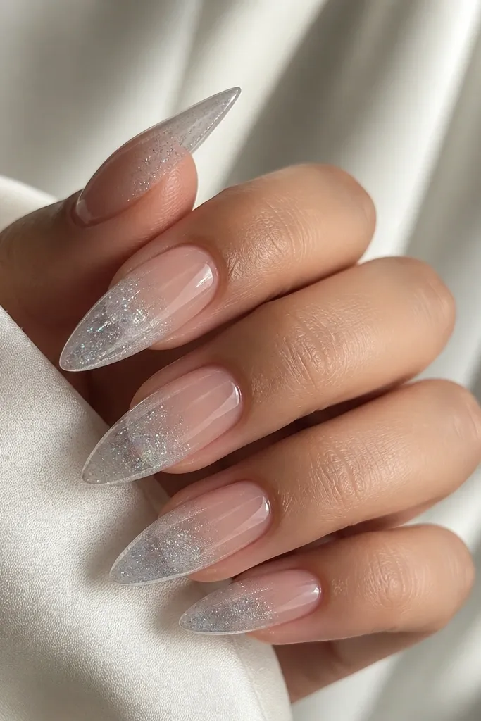 Almond nails with a clear base and negative space at the center. The tips fade from clear into icy silver shimmer, creating an ombré that stops before the cuticle. The nails look glossy and airy.