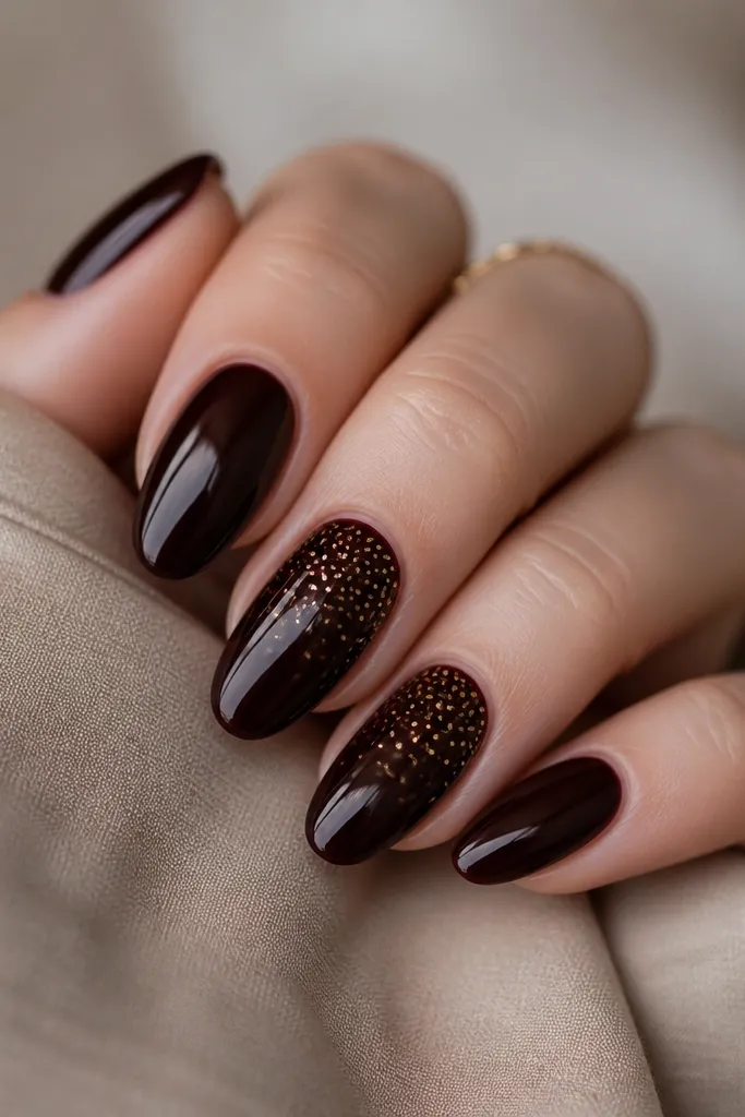 Glossy black cherry nails that look deep and reflective. On two accent nails, there are tiny gold dots scattered like snowfall, concentrated near the upper half and fading toward the tip.