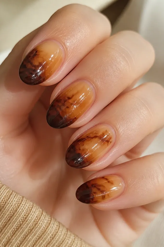 Close-up of short almond nails in warm honey beige. Each nail has irregular tortoise lines and caramel patches, with a darker espresso-brown tip fading into the mid-nail. Glossy topcoat catches light and shows a smooth, shell-like shine.
