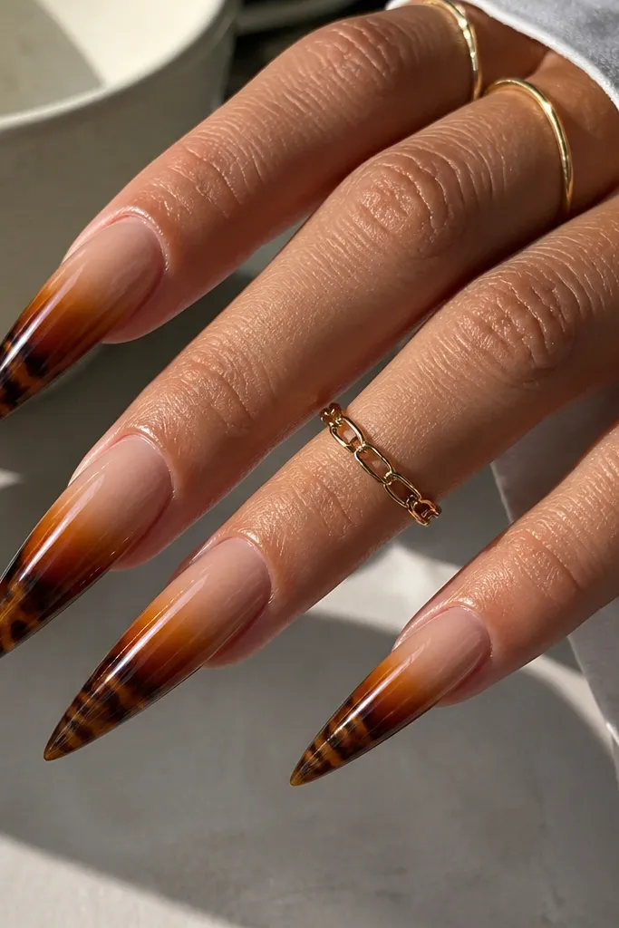 Long stiletto nails with a caramel glow gradient. The top near the cuticle is sheer nude, fading into caramel and darker tortoise patterns toward the tip. The tortoise includes irregular dark pockets and lighter streaks, all under a glossy finish.