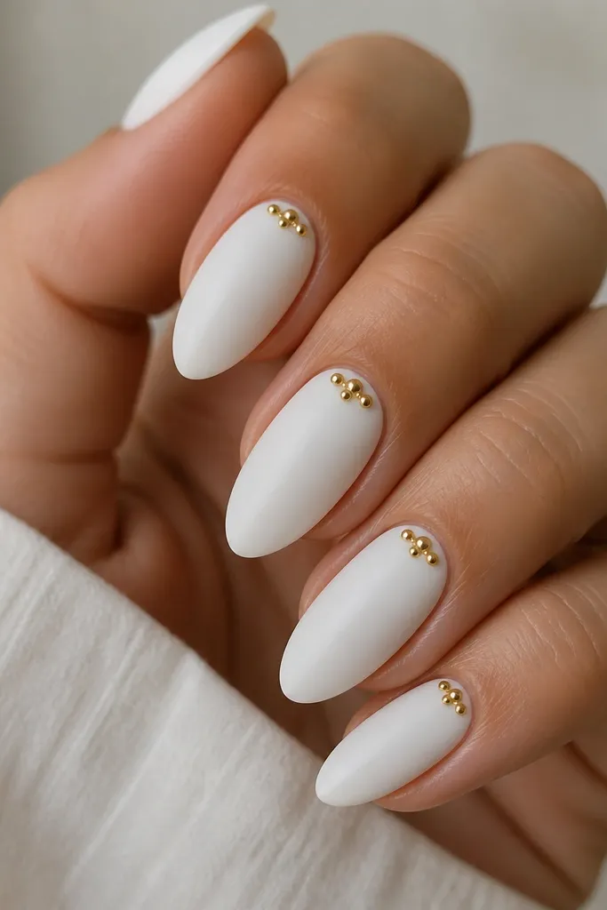 Medium almond nails with a soft satin-matte white finish. Near each cuticle, there are three tiny raised gold beads arranged in a small cluster, like a mini necklace. The beads are reflective and textured, standing out against the matte white. The nails are neat with crisp sidewalls.