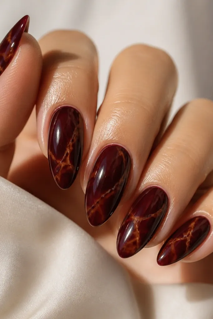 Five almond nails with a deep oxblood red base. Over each nail, thin amber and caramel tortoise veins run diagonally like marbled lines, with a glossy finish that reflects light.