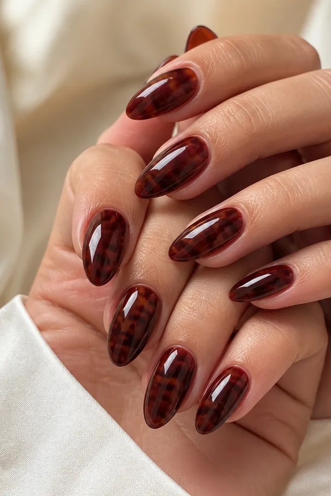 Medium-length almond nails with a wine-red base. The strongest tortoise pattern sits in the center, fading outward to a smoother red near the cuticle and side edges. Caramel-golden wisps sit between darker maroon patches, all under a high gloss top coat.