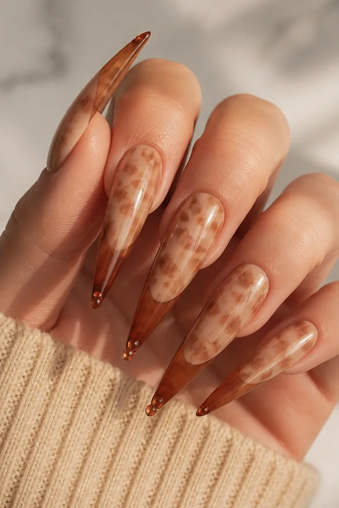 Very long stiletto nails. The base is a sheer nude with fine tortoise blotches in caramel and taupe. The French tips are stiletto-shaped and darker amber-brown. At the very tip of each nail, there are one or two small amber crystal dots placed like tiny jewels. Everything is glossy and reflective.