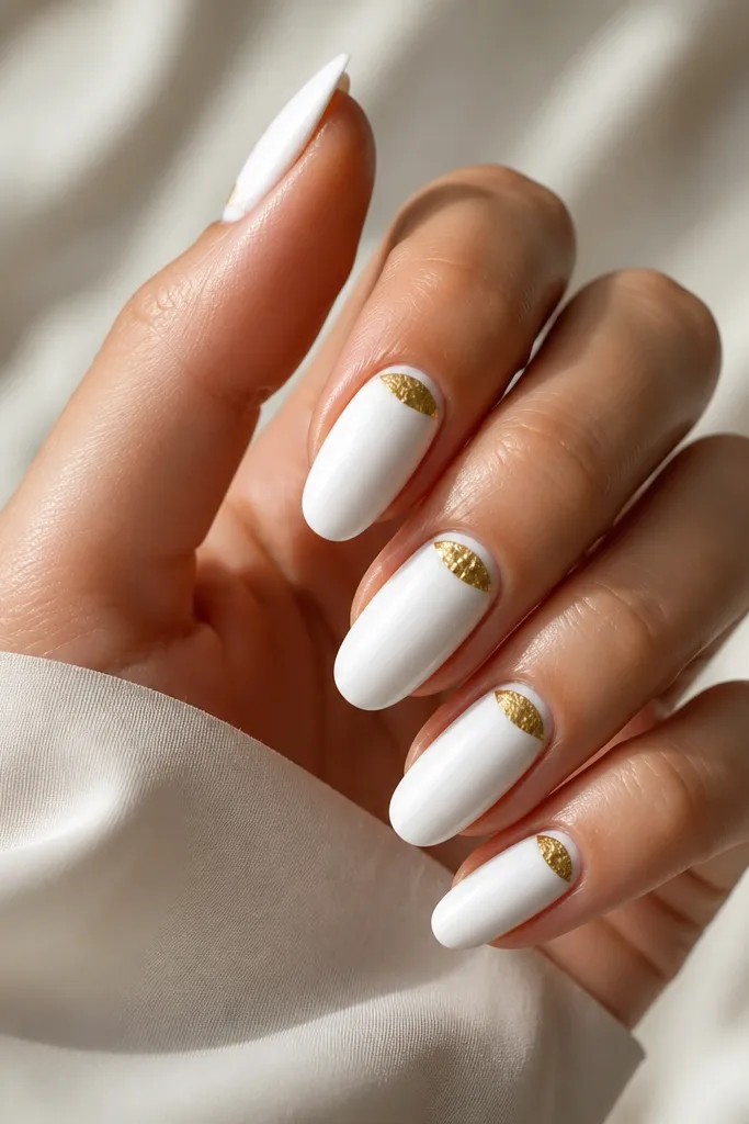 Long rounded nails painted bright glossy white. At the cuticle area of each nail is a crescent-shaped half-moon made of gold foil, with irregular foil texture. The foil sits right under the cuticle line and leaves a clean white border.