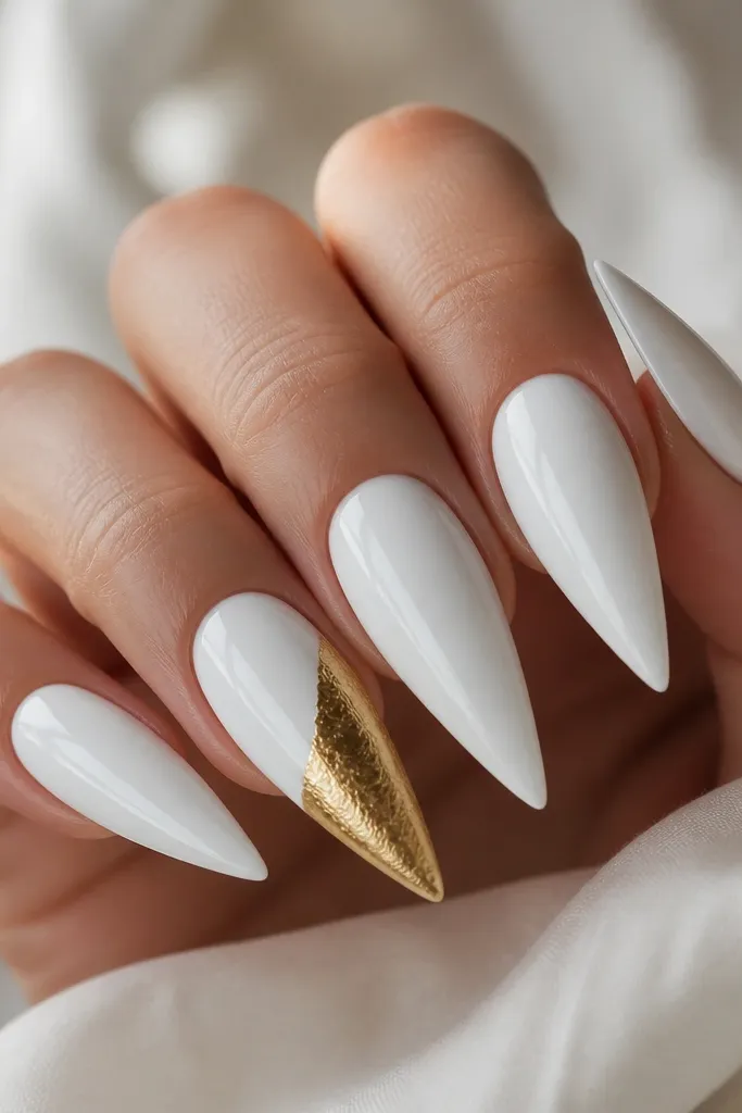 Five glossy white stiletto nails with a mirror shine. One accent nail has a gold foil half-moon that starts around the mid-nail and curves toward the tip, with irregular foil edges. The accent nail has a slightly textured foil sparkle, while the other nails are smooth and solid.