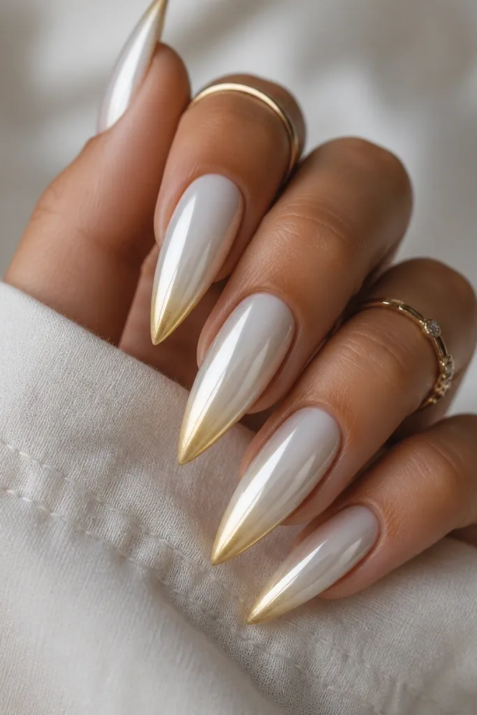 White stiletto nails with a chrome-like sheen. The top half looks silvery-white and fades into a warm gold at the very tip. The transition is smooth and blended, with a gradient that looks like light shifting across the nail.