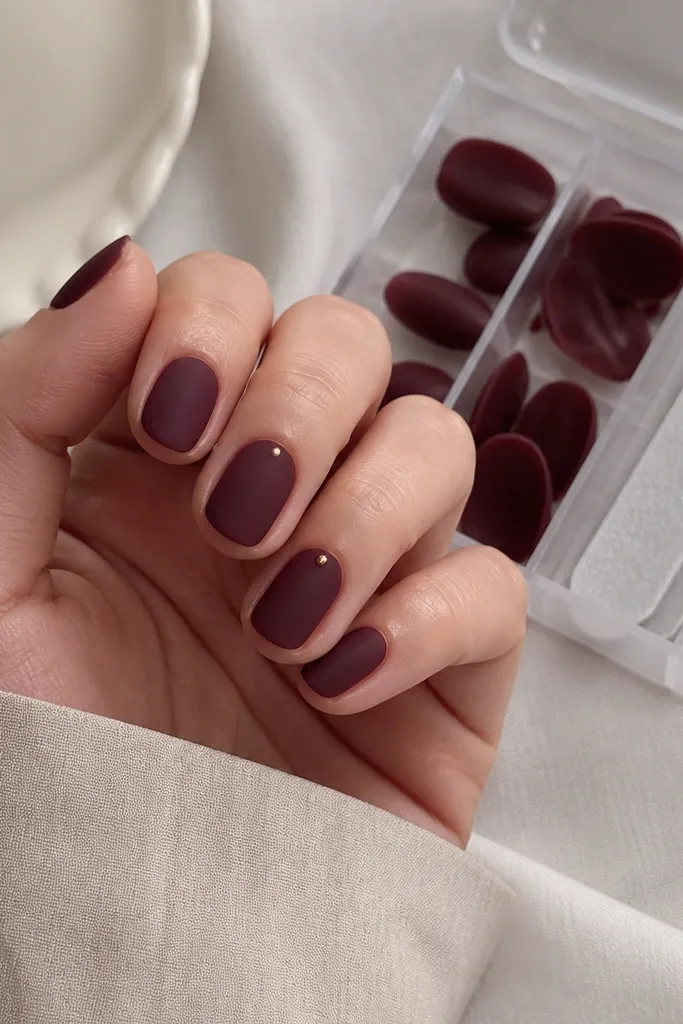 Short oval nails in deep burgundy. The finish looks velvety matte, not shiny. One nail has a tiny gold dot at the cuticle. A clear tip case is open in the background with spare tips and a mini file.