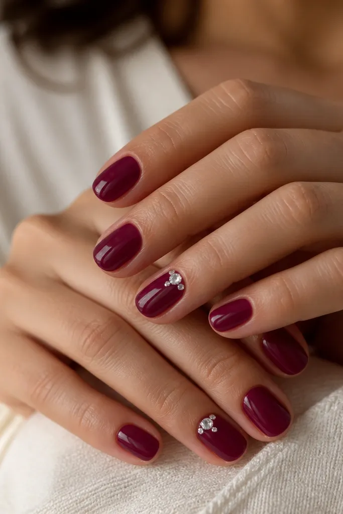 Short oval nails painted in a glossy berry jelly polish. On two accent nails, there is a tiny cluster of clear crystals placed near the cuticle, with one crystal slightly larger.