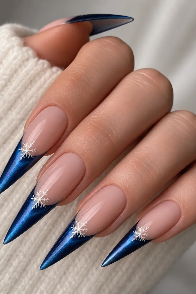 Long stiletto nails with a nude base. French tips are ink blue cat eye with a bright streak running down the center. Tiny white micro snowflake decals sit near the cuticle side of the tip, and the rest is glossy.