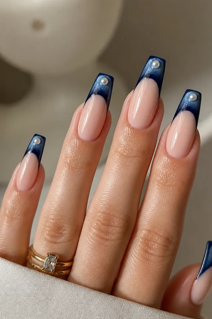 Long coffin nails with a nude base. French tips are deep navy cat eye with a bright streak. In the center of each tip, there's a small glazed pearl bead effect, like a drop of glossy pearl gel, and the nails are shiny.