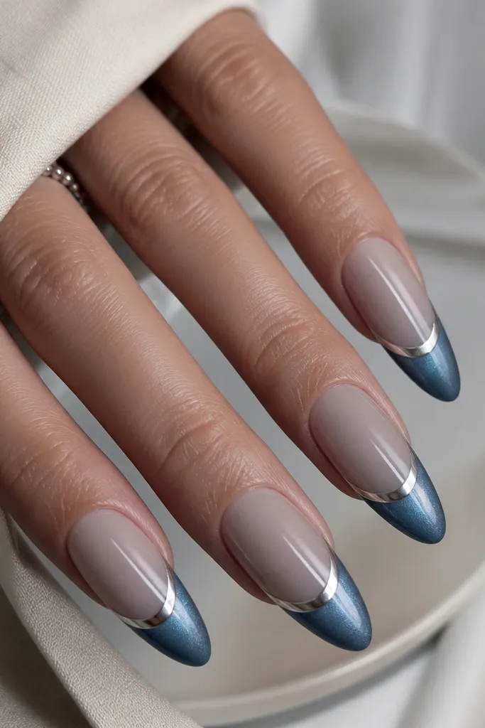 Long almond nails with a milky smoke-gray nude base. The French tips are steel blue cat eye, pulled into a sharp arc near the cuticle line. A thin chrome halo ring sits at the base of the tips, like a rim of silver mirror. The cat eye glow shifts from slate to blue as the light hits, with a soft smoky gradient into the base.
