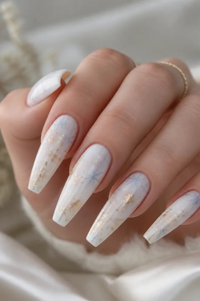 Long coffin nails on a milky white marble base. There are small icy blue starburst shapes near the cuticle and along one side, with scattered gold foil flecks concentrated near the center.