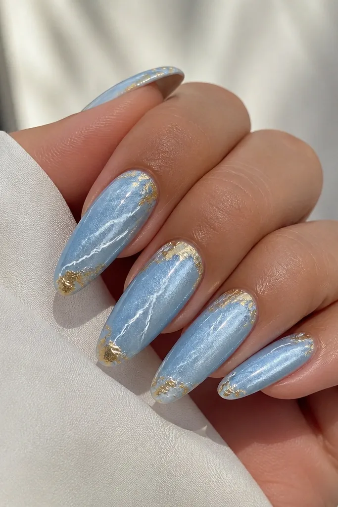 Long almond nails painted icy blue. White lightning bolt lines run diagonally across each nail, and gold foil corners sit near the cuticle and near the tip.