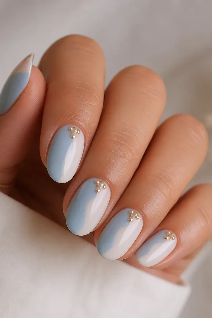 Medium oval nails split into two halves by a curved swirl. One side is pale blue, the other is milky white with a soft swirl edge. Near the cuticle sits a tiny gold bulb cluster with one or two white dots.