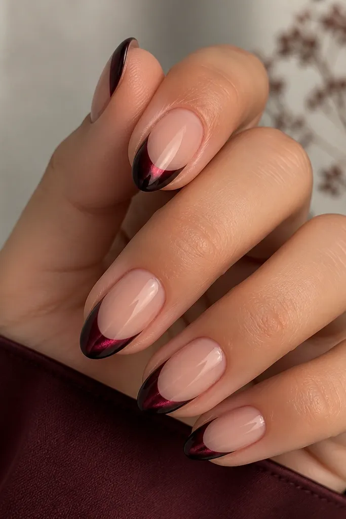 Short-to-medium almond nails with a sheer nude base. French tips are black cherry chrome cat-eye, deep maroon with a bright wine-red stripe. The stripe shifts toward dark plum at certain angles.