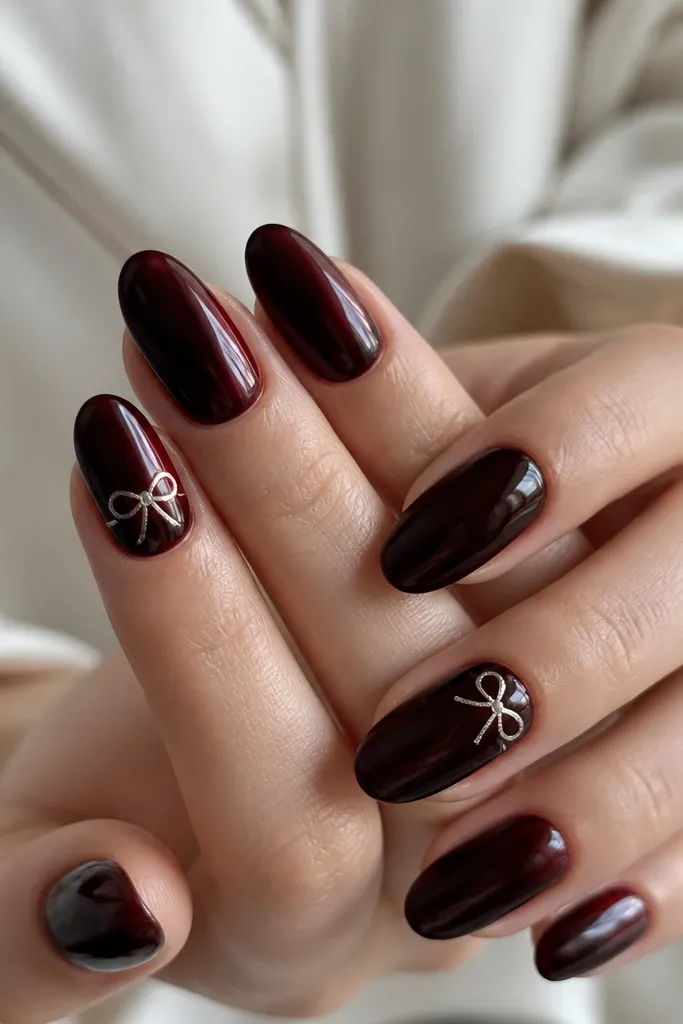 Glossy black cherry nails with a deep wine sheen. Two accent nails have a tiny silver bow drawn near the cuticle, with thin lines and small dot centers.