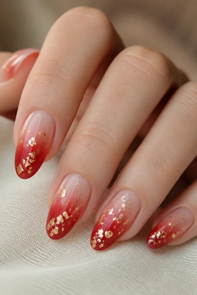 Medium oval nails with a smooth red-to-nude gradient; gold leaf flecks float near the center and tip, scattered lightly across each nail.