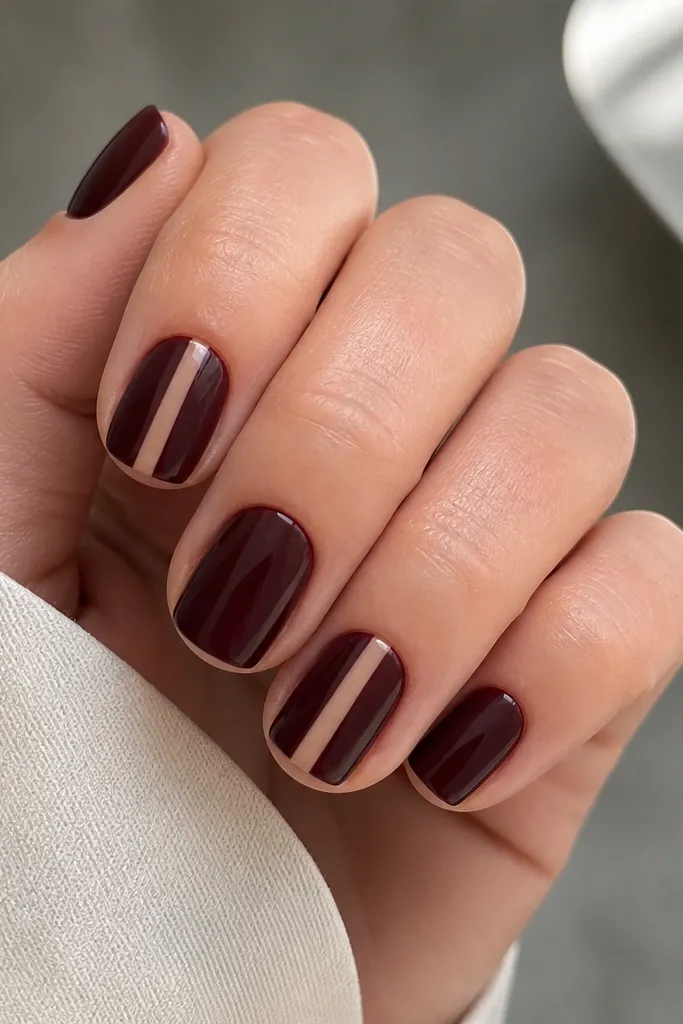 Short nails painted glossy burgundy. On one nail (or two), there is a vertical nude panel near the center that peeks through like a window. The panel is narrow, with clean edges, and the burgundy wraps around it. The rest of the nails are solid burgundy.