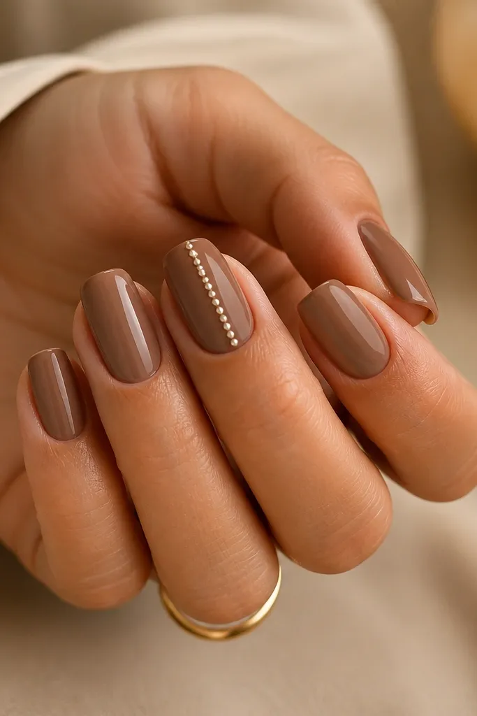 Mocha nude square nails in a warm beige-brown base. On the middle finger, there's a vertical pearl stripe running from near the cuticle to the top, made of tiny dots aligned in a line. The nails are glossy and the pearl stripe is centered.