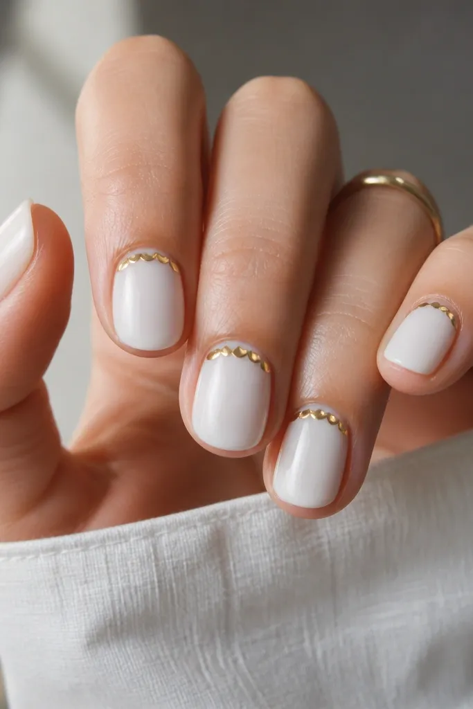 Milky white nails with a series of small gold scallops along the cuticle line, like tiny arches, glossy top coat.