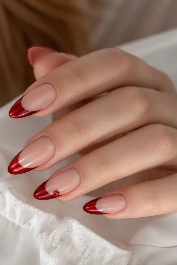 Almond nails with a nude base. The french tips are red cat eye with a strong vertical stripe. One tiny red rhinestone sits near the french curve on the ring finger, with clear gem sparkle on top.