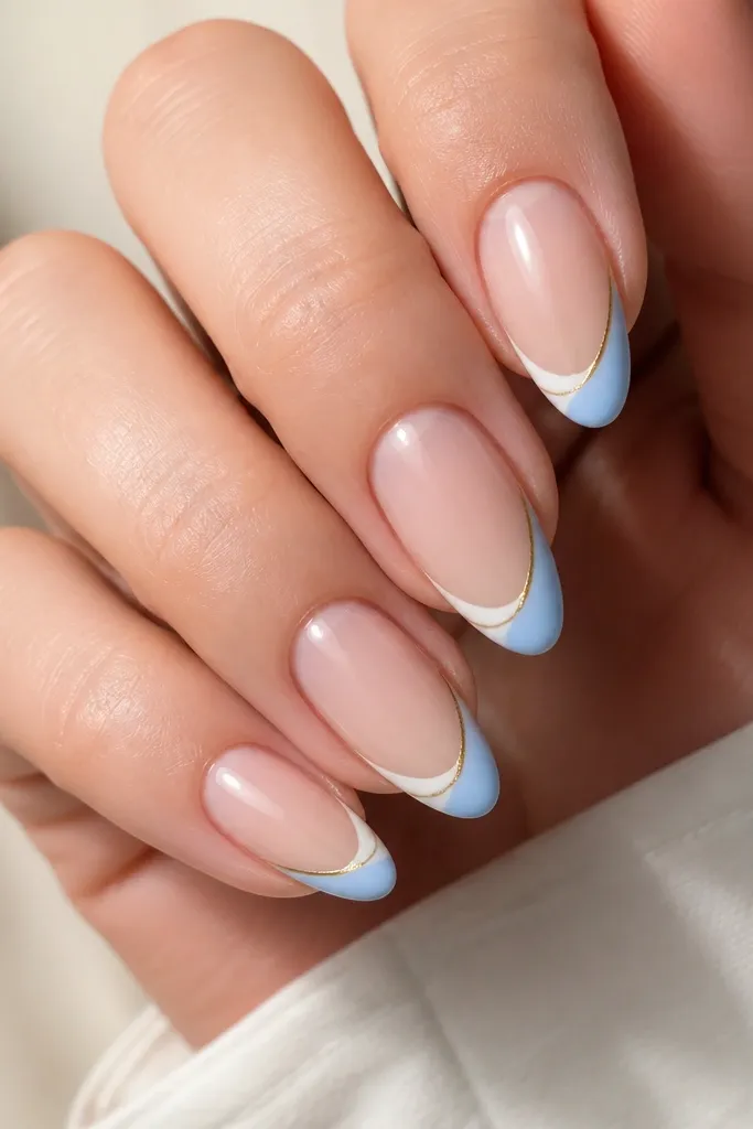 Close-up of almond nails on a light skin tone. The base is sheer nude. The tips are painted in bright blue-white french, and a thin 14k-style gold stripe runs parallel just inside the white edge. One nail shows a tiny gold dot at the corner.