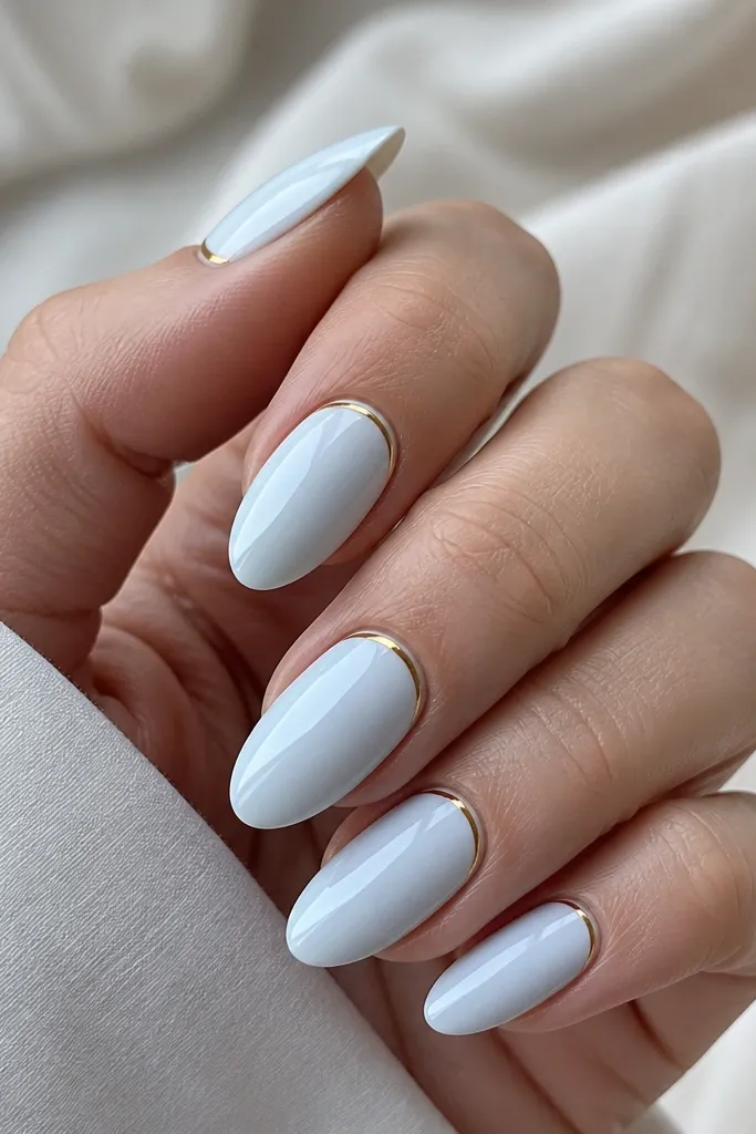 Almond nails with a blue-white opaque base. At the cuticle, a thin gold half-moon sits exactly along the natural nail curve. The rest of the nail is solid blue-white with a glossy finish.