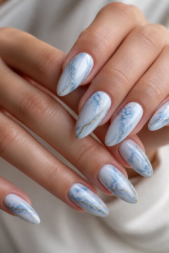 Almond nails with a blue-white marble pattern. Fine darker blue-gray veins twist through the white. Thin gold lines trace one side of each vein on alternating nails.