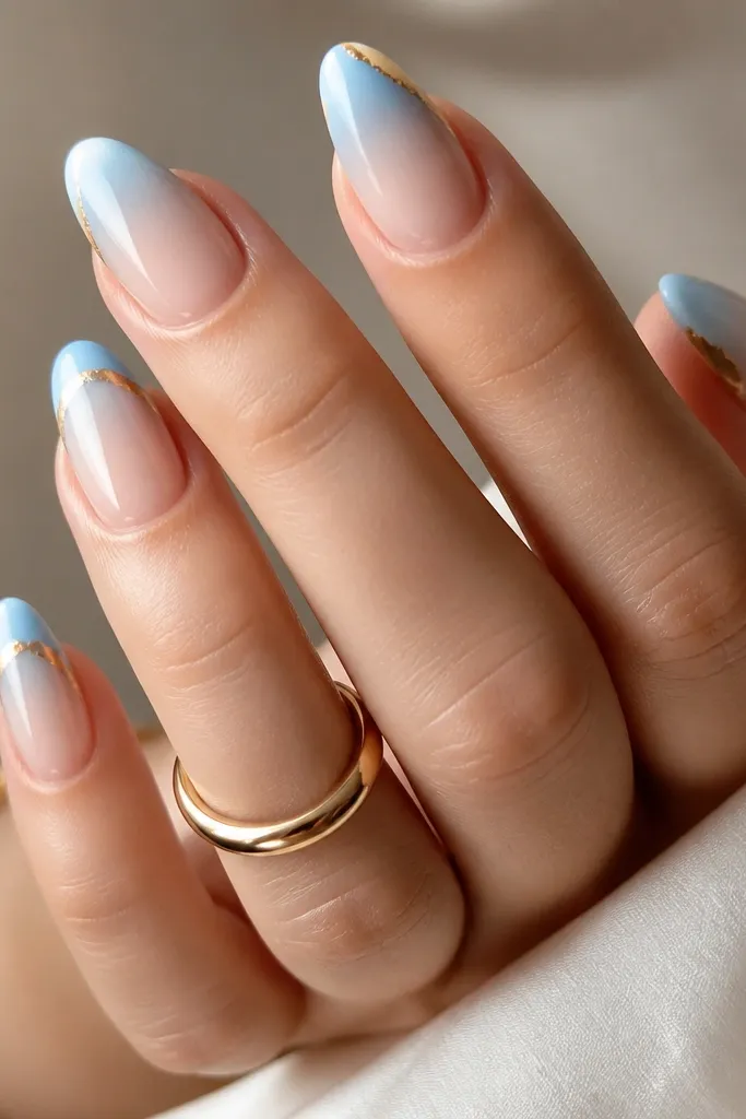 Almond nails with a blue-white-to-sheer nude ombre. Near the tip, a gold foil edge wraps across the outer rim like a metallic border.