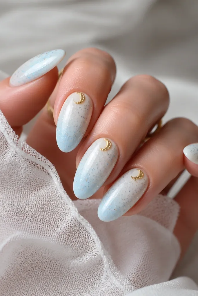 Almond nails painted blue-white. Random tiny dots in light blue and silver scatter like a galaxy. A gold crescent sits near the cuticle on each nail.