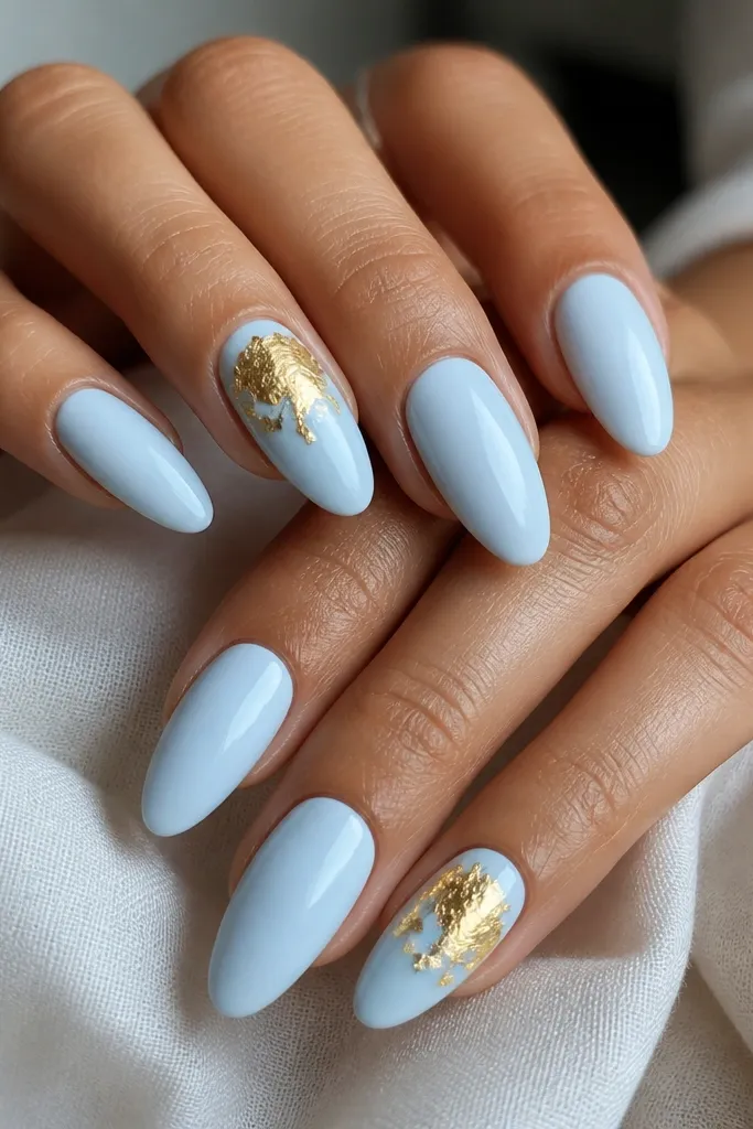 Almond nails fully covered in glossy blue-white. On two accent nails, gold leaf pieces sit like irregular patches near the top third, with a few edges lifted slightly under the shine.