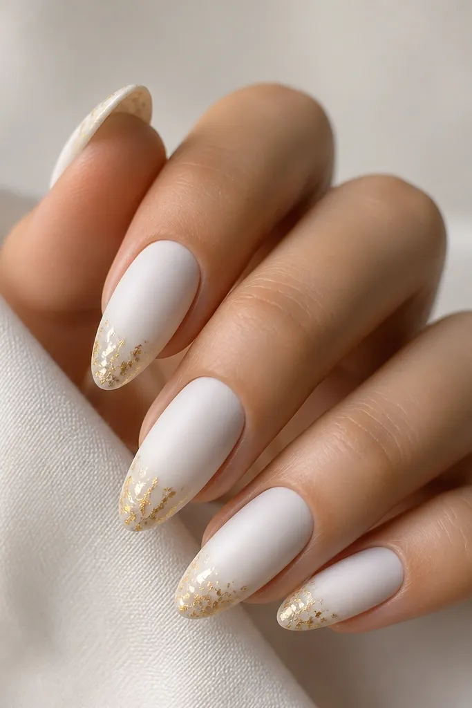Five almond nails with matte white polish. At the tip area there are thin, flaky gold leaf pieces clustered lightly along the edge. The cuticle stays plain matte white. Matte texture is visible, not glossy.