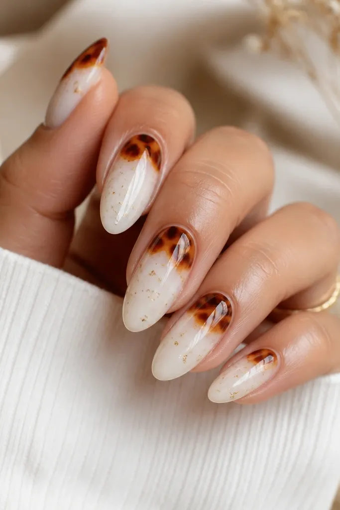 Almond nails with milky white base. Warm tortoise patches sit on the tips and upper third, and tiny gold flecks are scattered sparingly across the white. Nails are glossy and catch light.