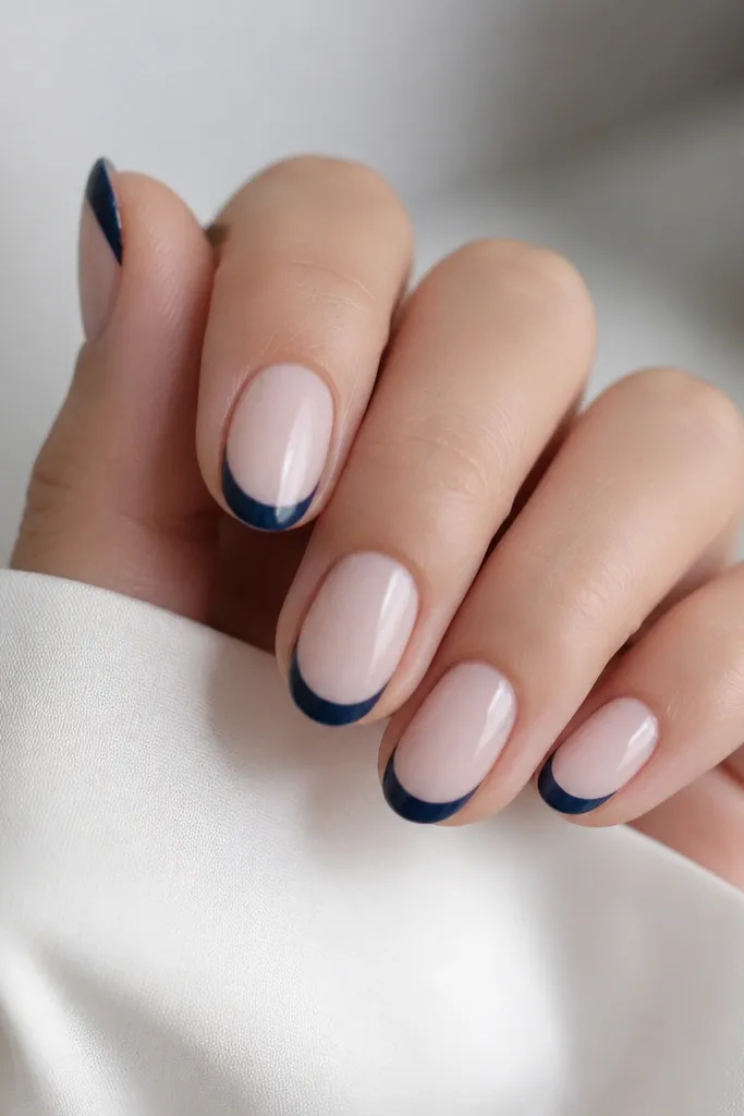 Short oval nails with a milky base and a navy blue French tip shaped like a smooth smile curve; glossy top coat, no other patterns.