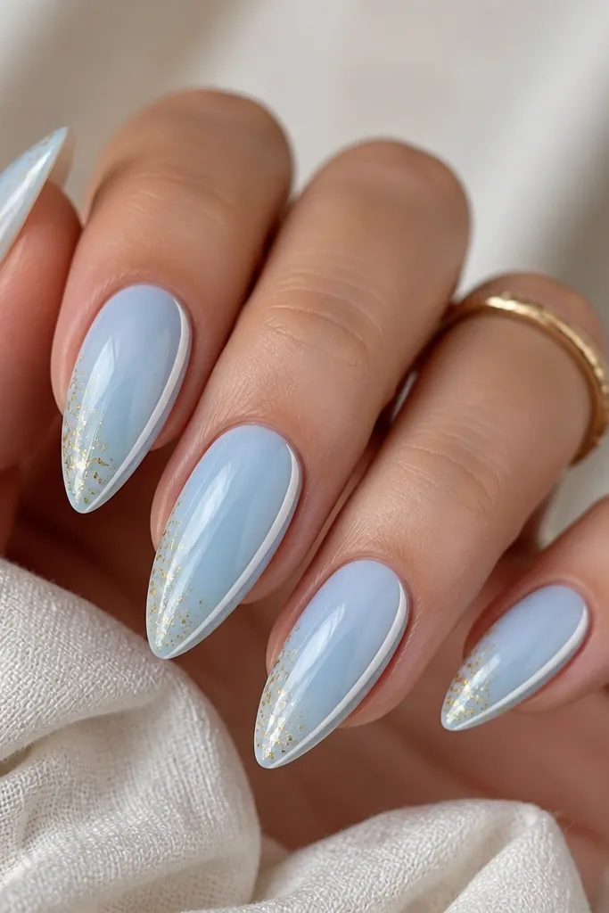 Long almond nails with a milky baby blue base. Only one side of the tip has a thin white French stripe, and scattered gold foil speckles sit near the tip on the opposite side. Everything is glossy, with small glints from the foil.