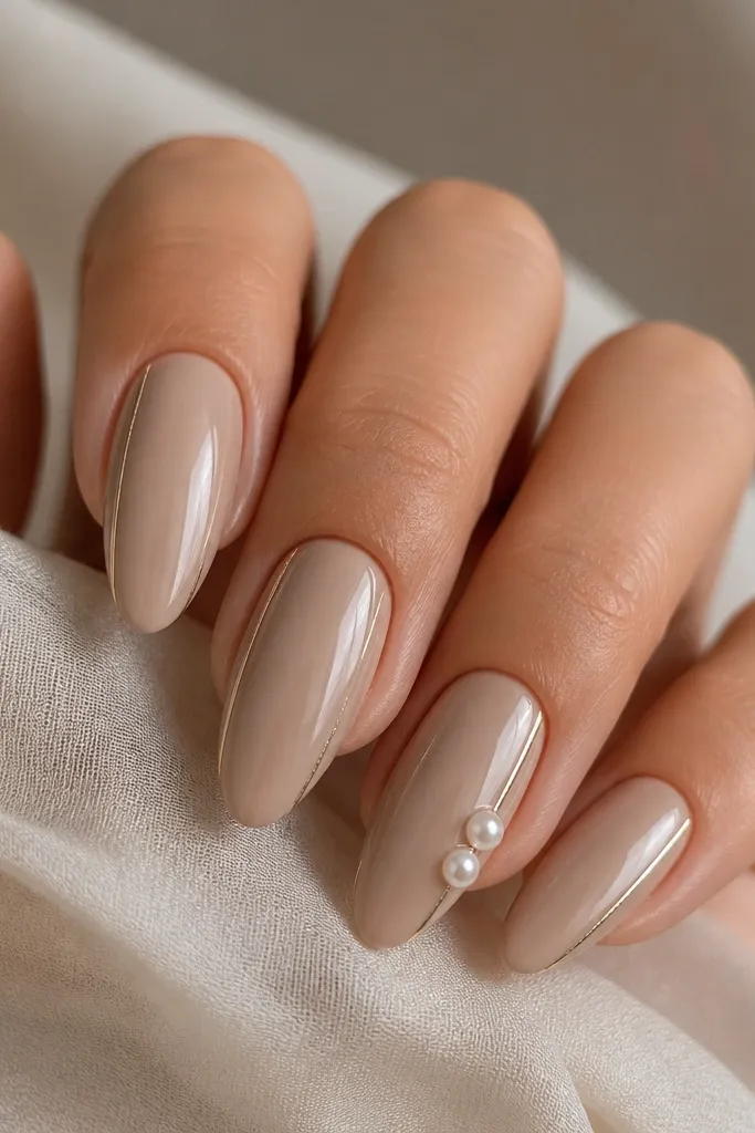 Medium almond nails in a champagne nude glossy base. On one accent nail, there's a thin metallic side bar near the outer edge and two small pearl studs placed near the bar. Other nails have only the metallic bar without pearls.