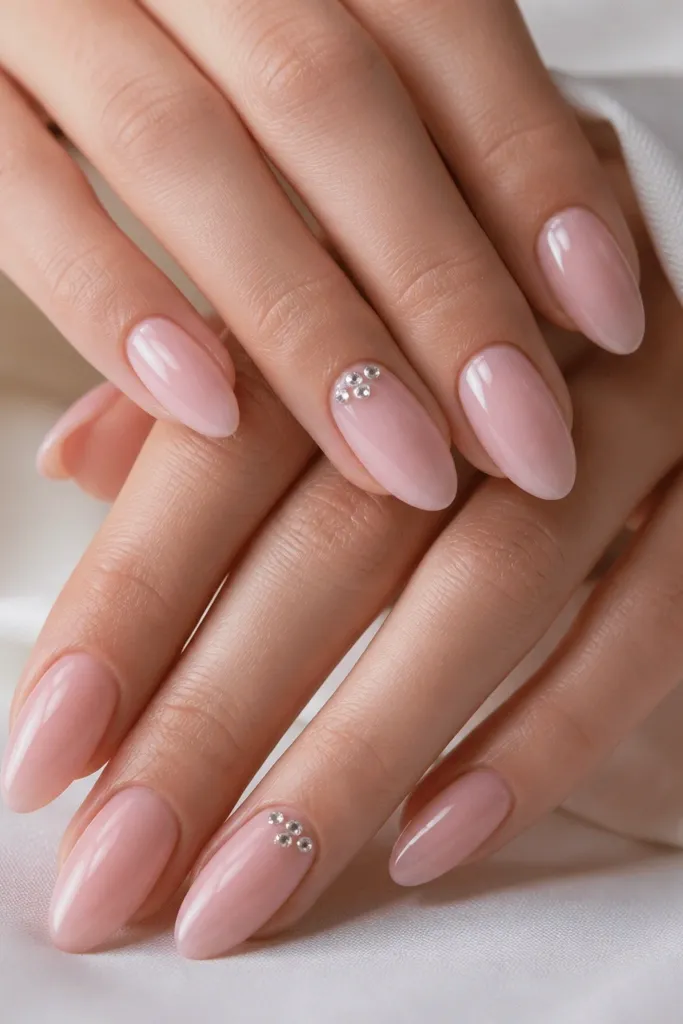 Almond nails in a soft blush pink. On two nails, there's a tiny cluster of clear micro gems near the cuticle - three to five small stones arranged like a small constellation. The nails are glossy with no bulk around the stones.