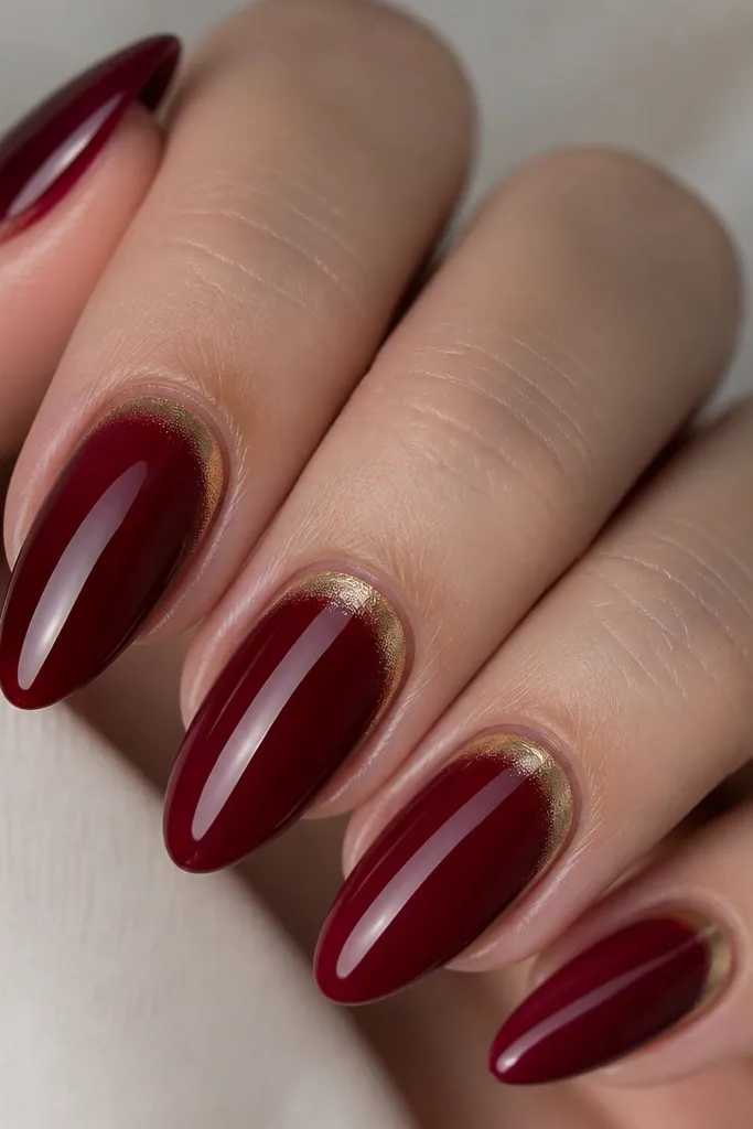 Close-up of almond nails painted in a deep cherry red with a high-gloss finish. The cuticle area has a thin, pale-gold chrome ring that fades into the red, forming a soft halo. The tips are clean and smooth with no glitter across the nail.