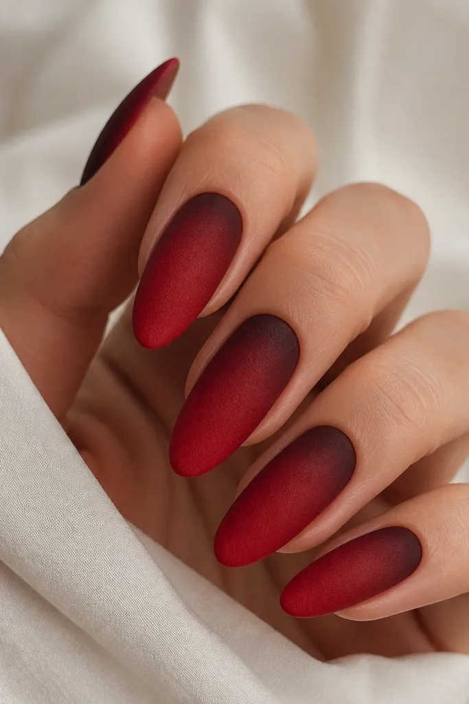 Almond nails painted in a matte-to-velvet finish red. The cuticle area is a deeper burgundy, fading into a brighter red toward the middle and tip. No glitter, just a soft, fuzzy-looking velvet surface under light.