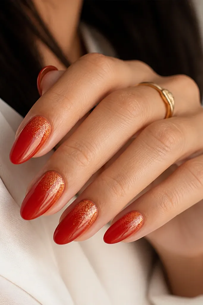 Almond nails painted in a warm tomato red glossy base. Gold micro speckles appear near the cuticle and fade toward the middle, leaving the tips mostly clean red. The speckles are fine and evenly scattered, not chunky.