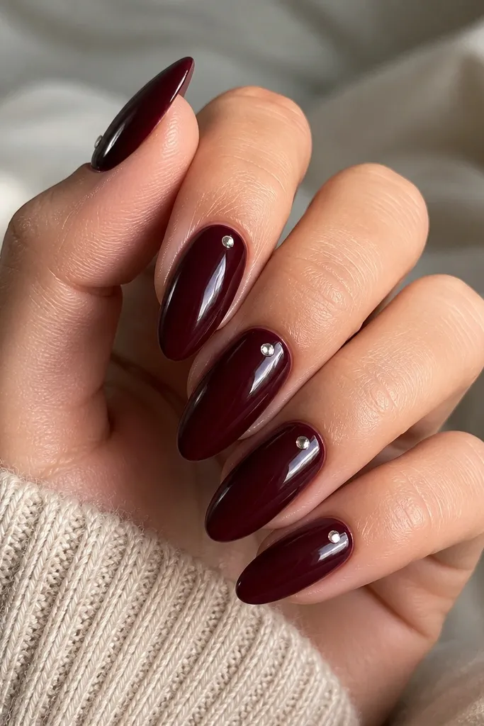 Almond nails in deep burgundy glossy polish. On each nail, a single small clear rhinestone sits slightly off-center along the side near the cuticle, catching light. The rest of the nail is plain burgundy, no other gems.