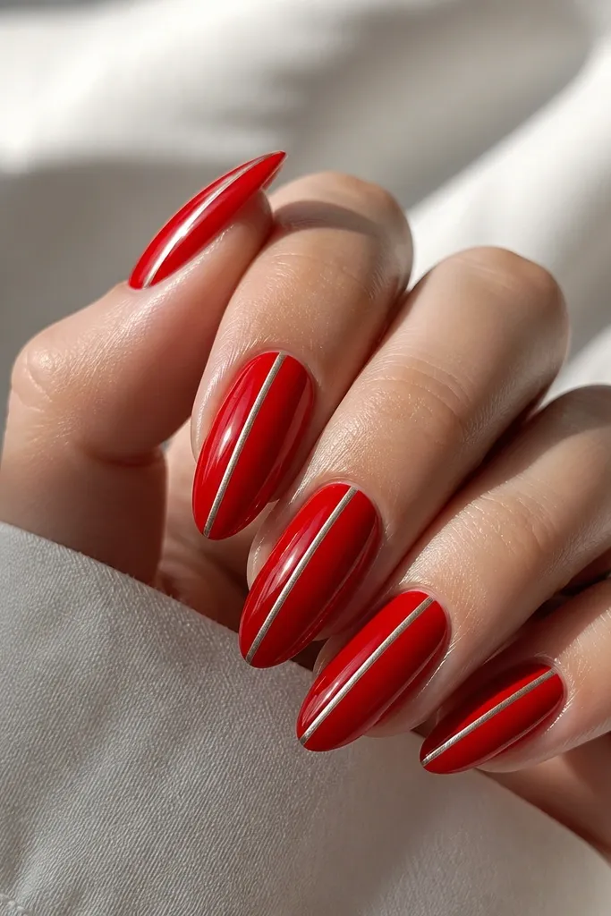Glossy red almond nails with a single ultra-thin chrome stripe running vertically near one sidewall. The stripe is silver-chrome and catches light as a narrow line. No other decoration, just clean red and one reflective detail.