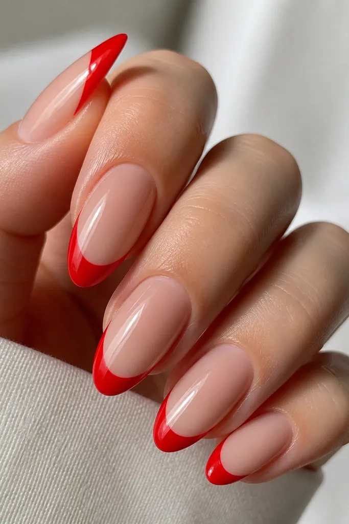 Close-up of almond nails on a hand with a glassy sheer nude base. The french tips are bright true red with a crisp curved smile line, glossy top coat, no gems or extra art.
