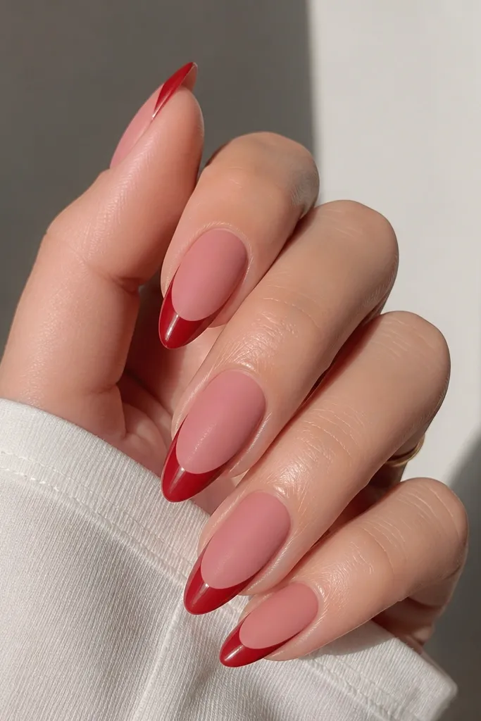 Almond nails with a matte dusty pink base. The french tips are glossy bright red with a clean curved line and a sharp almond outline; the tips reflect light while the base stays velvety.