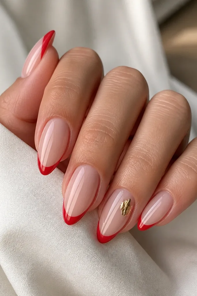Almond nails with sheer nude base and glossy red french tips. On the ring finger, a small irregular piece of gold foil sits near the cuticle or side, with no other glitter; all nails are glossy.